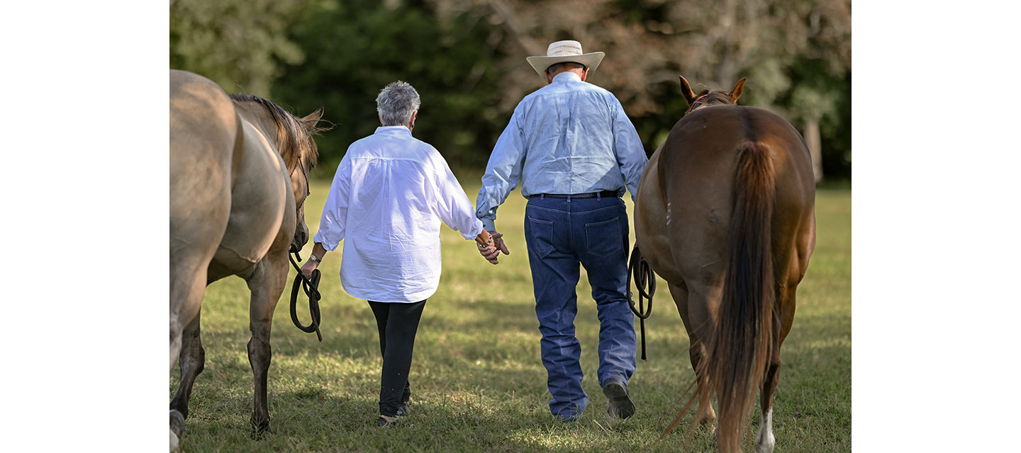 An older couple walks hand-in-hand through a pasture, leading two horses, with their backs turned to the camera.